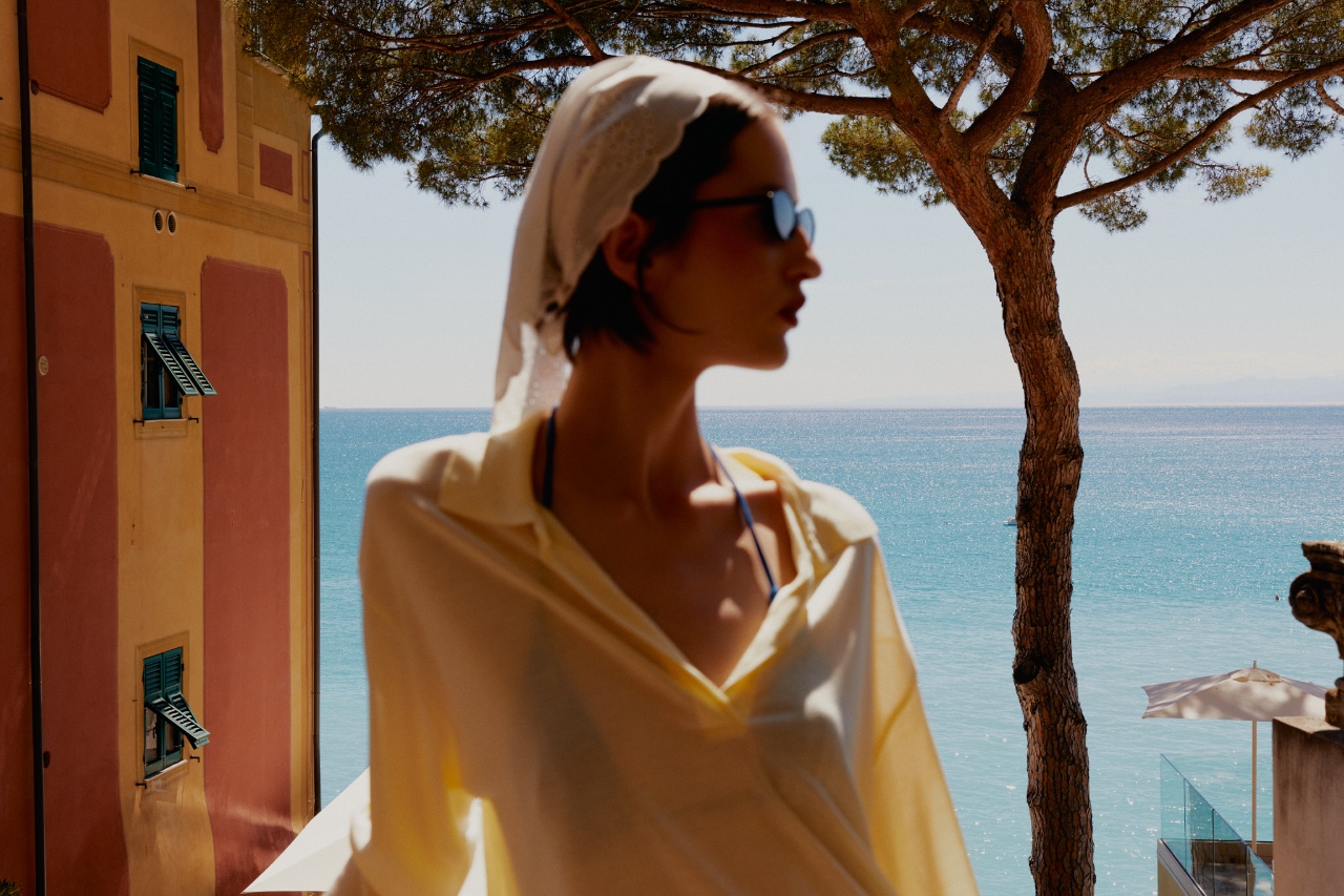 Woman in a cream headscarf on a sunlit Mediterranean terrace above turquoise sea