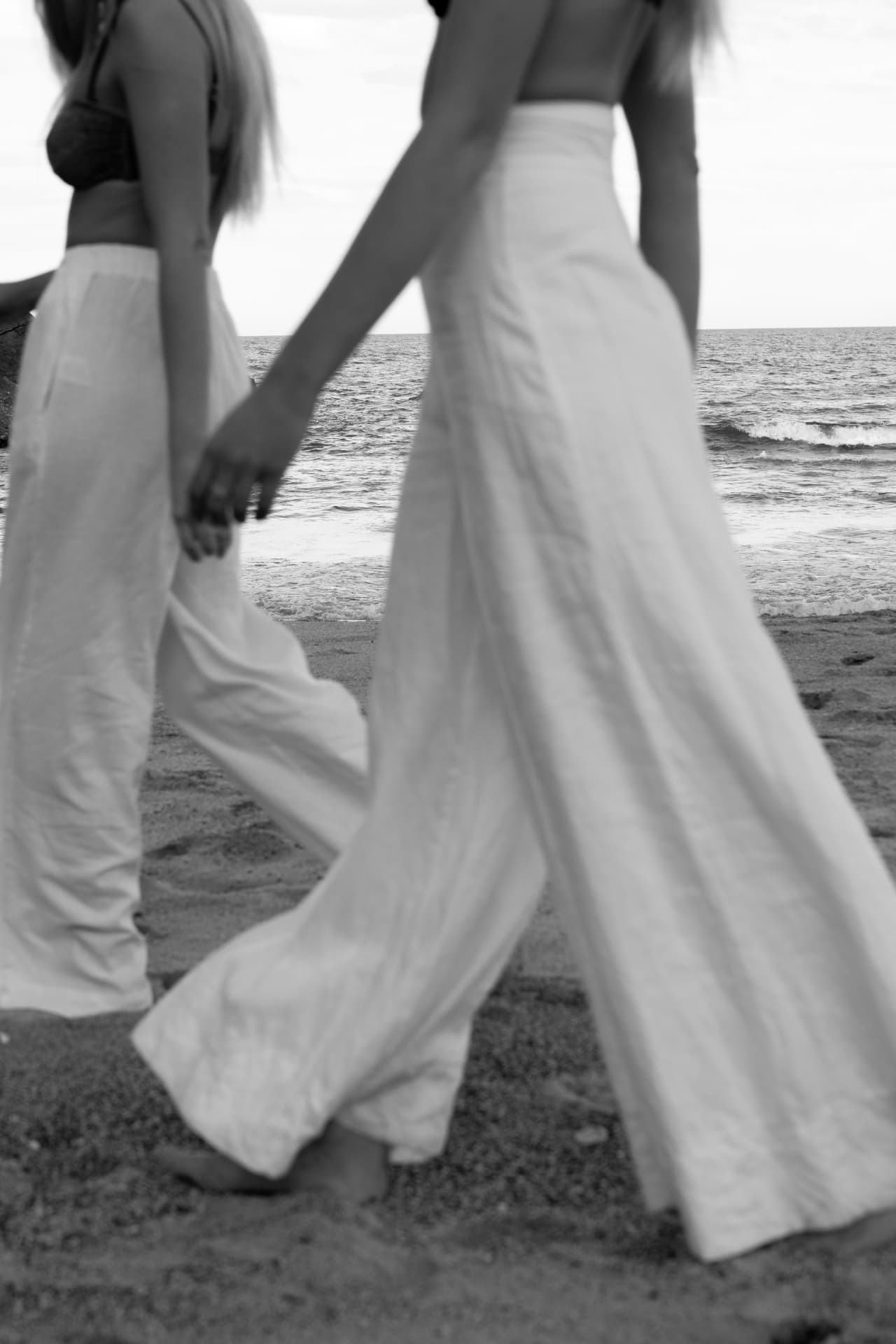 Two women in flowing white trousers holding hands at the water's edge at dusk
