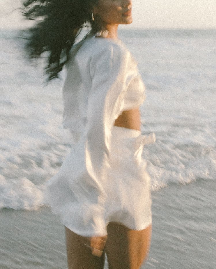 Woman in a white dress wading through ocean waves at golden hour