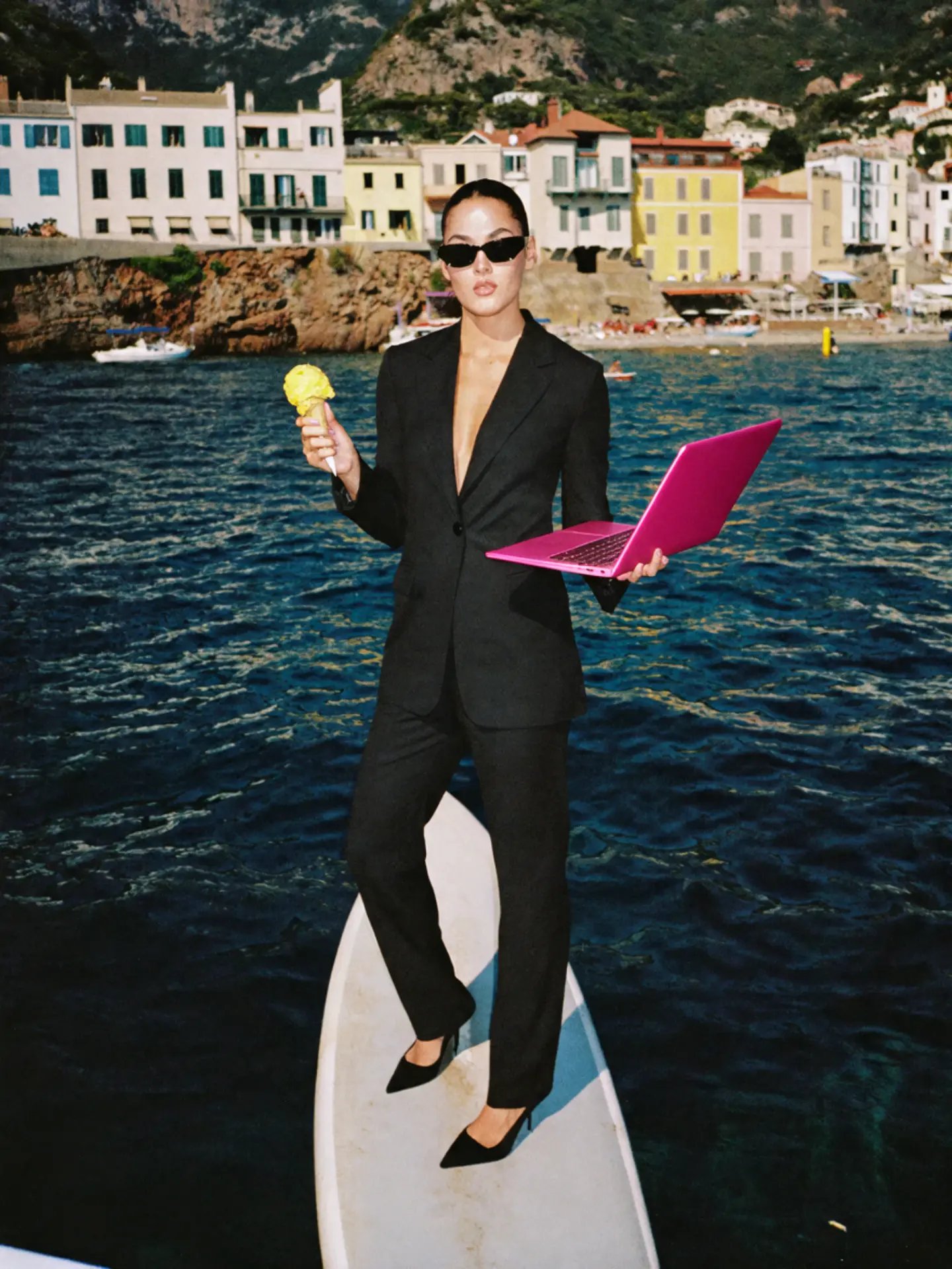 A woman in a black suit and sunglasses eating a yellow ice cream while holding a hot pink laptop, standing on a surfboard on the sea with a coastal village on cliffs behind her