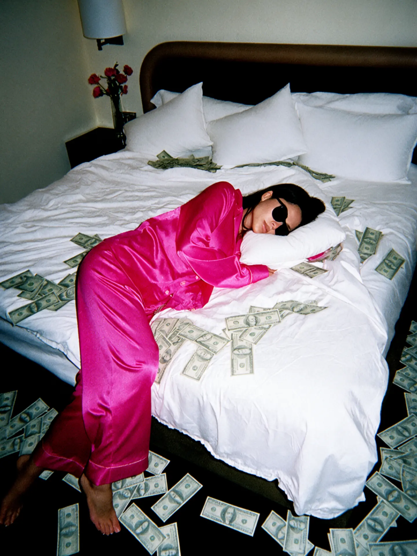 Woman in pink satin pyjamas lying on a bed surrounded by scattered cash