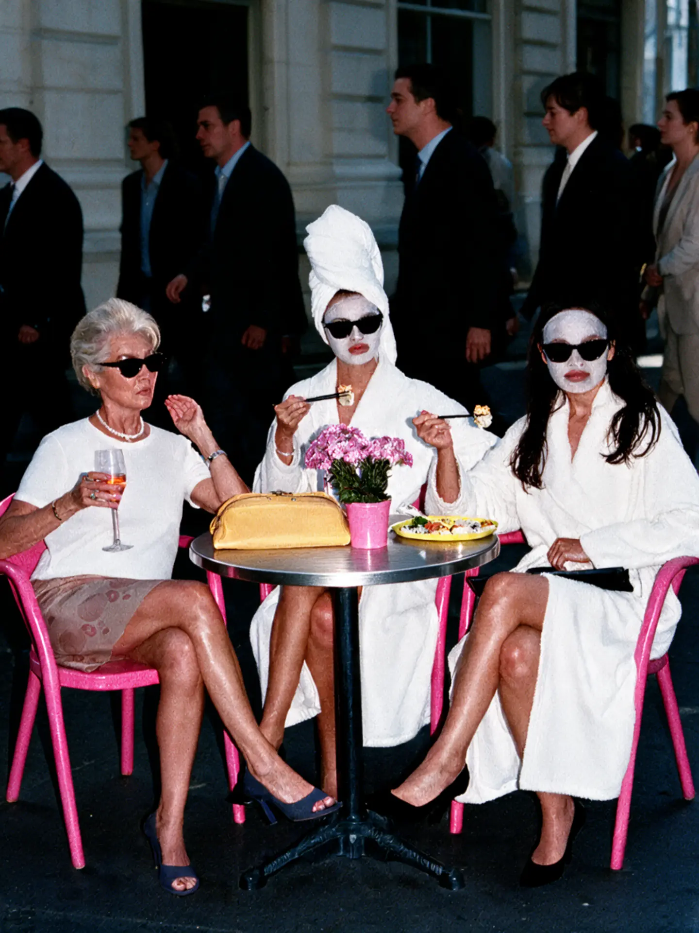 Three women in spa robes and sunglasses enjoying food at a city street table, joined by an older woman with a drink